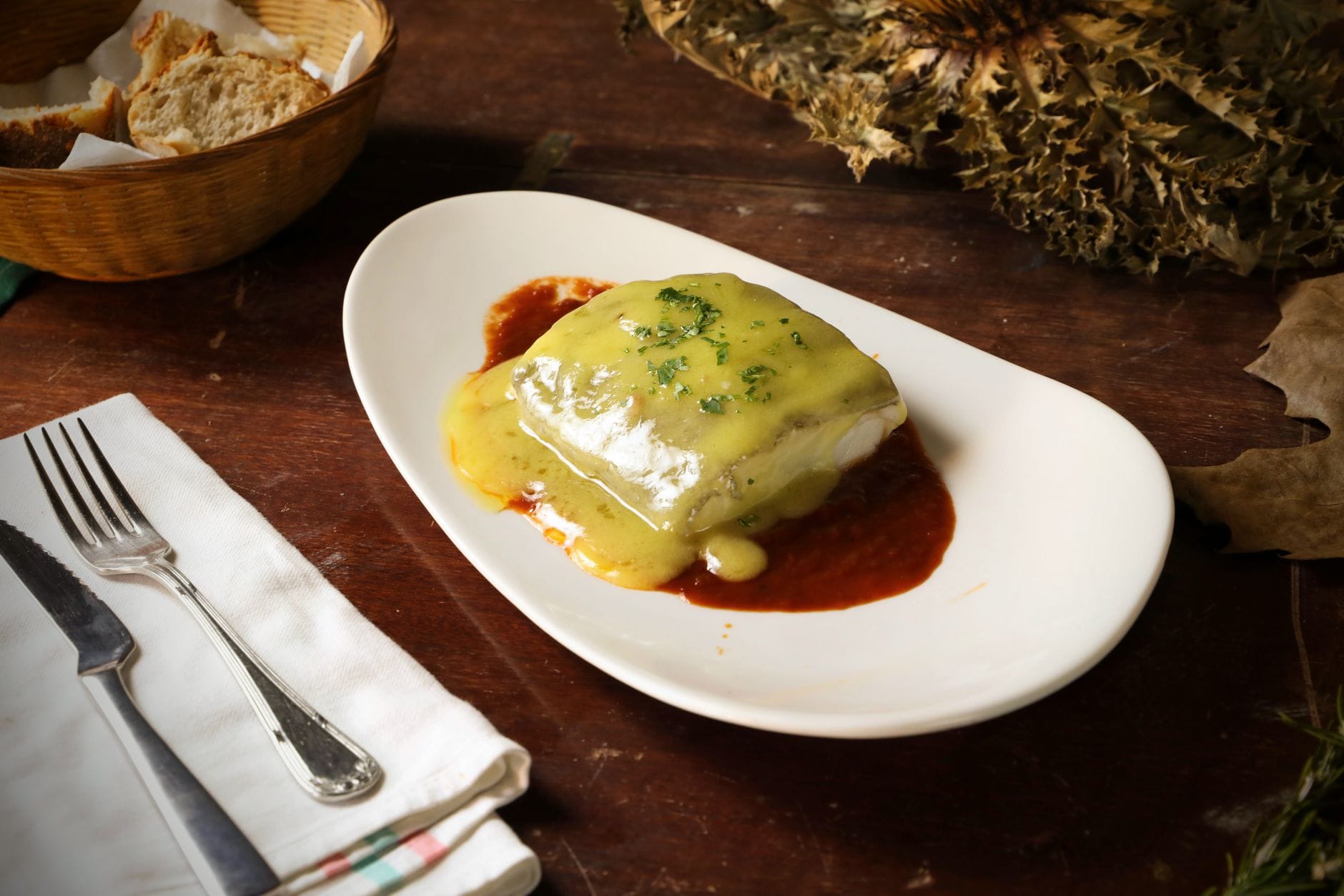 Un piatto di bacalao al pil-pil con una salsa densa ed erbe aromatiche su un tavolo di legno rustico, servito con pane e posate.