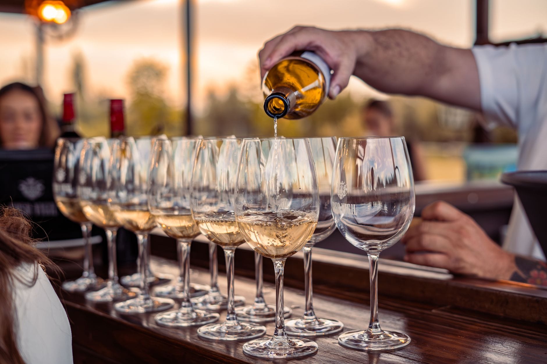 Una fila di bicchieri da vino su un bancone di legno, con qualcuno che versa vino bianco, persone sfocate sullo sfondo.