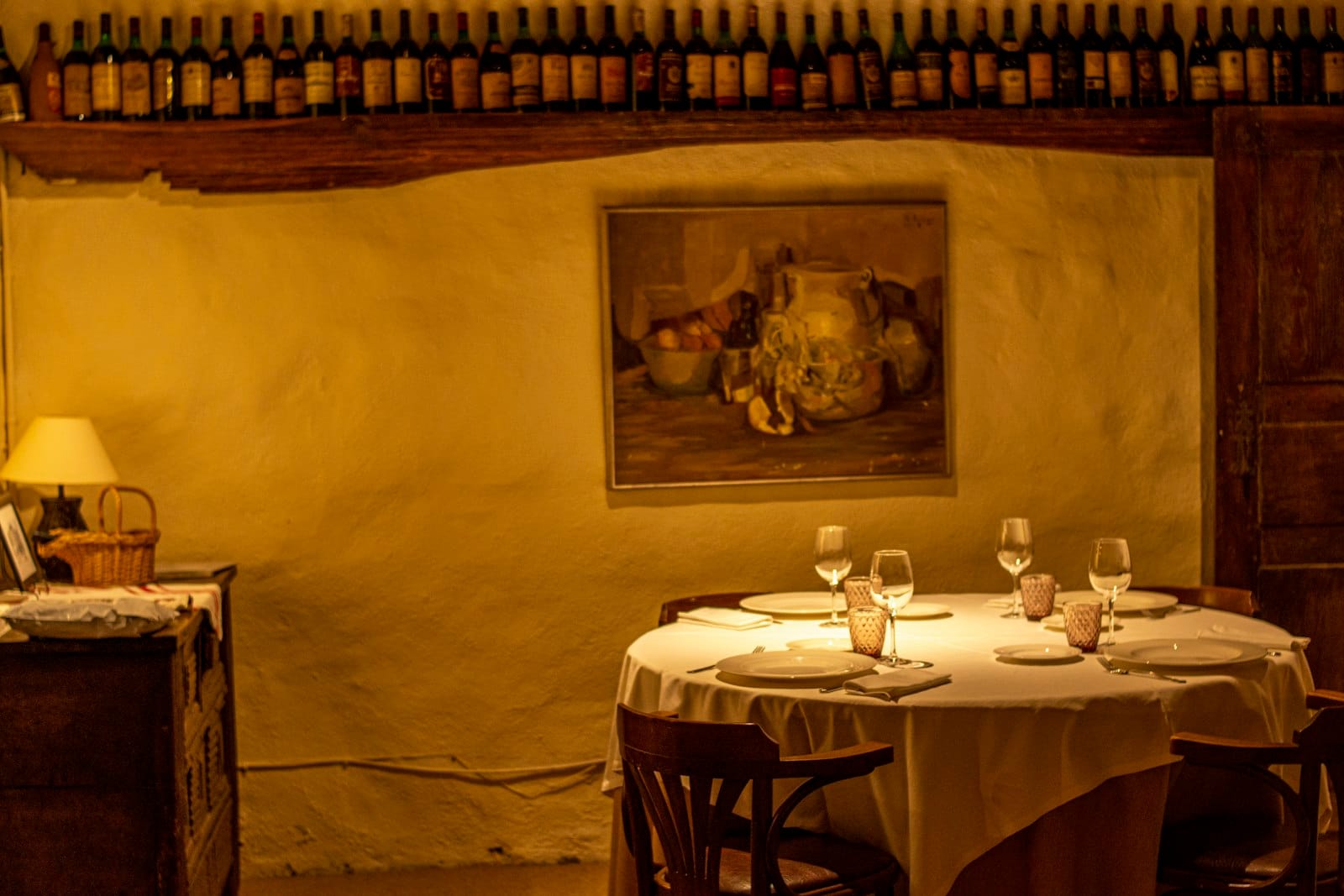 Warmly lit rustic Basque restaurant interior with round table set for four, wine glasses, and a shelf of wine bottles, evoking local food culture in Bilbao.