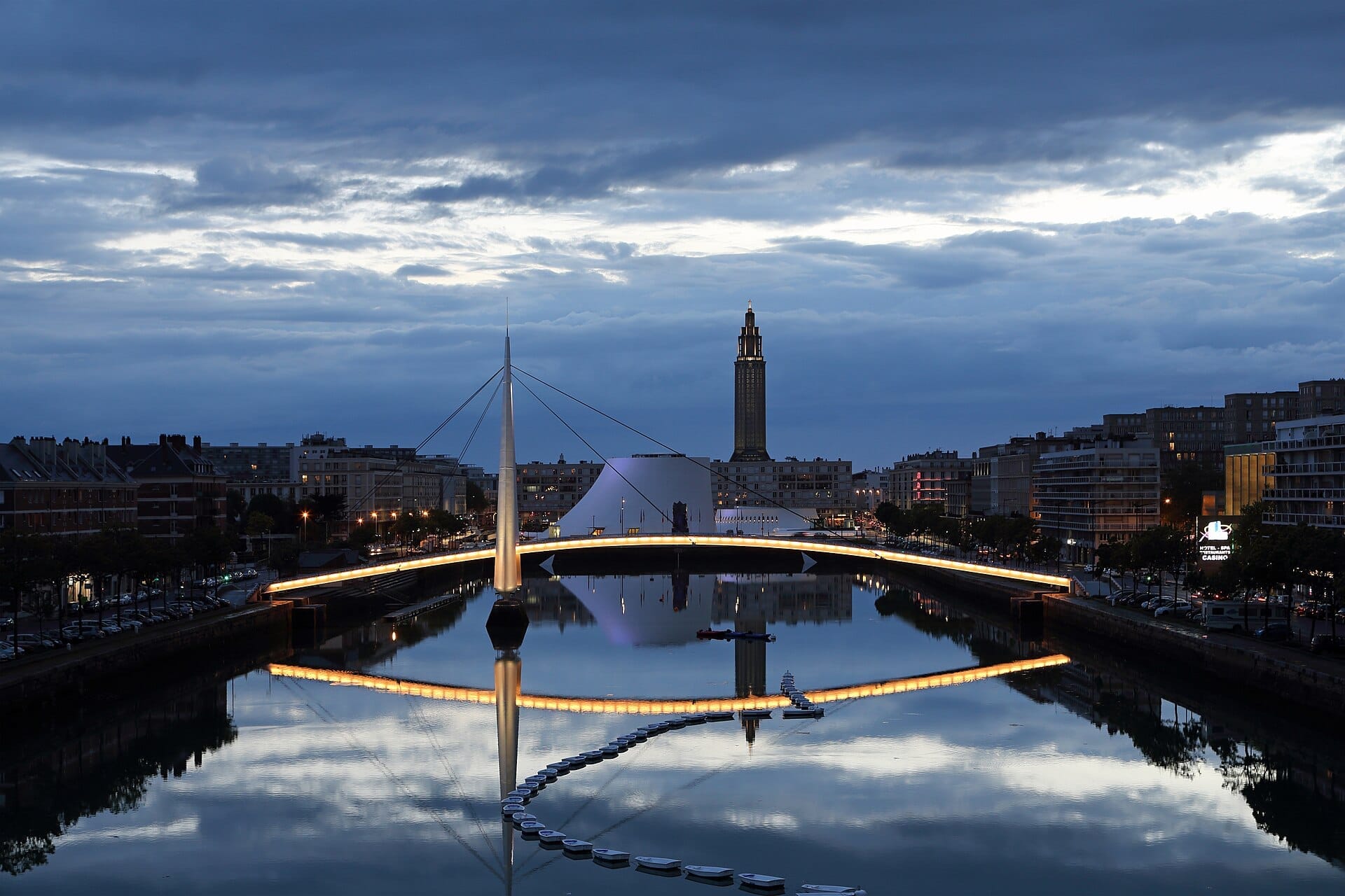 Vista serale del Bassin du Commerce a Le Havre, con ponti moderni, edifici sul lungomare e il riflesso della torre sotto un cielo nuvoloso spettacolare.
