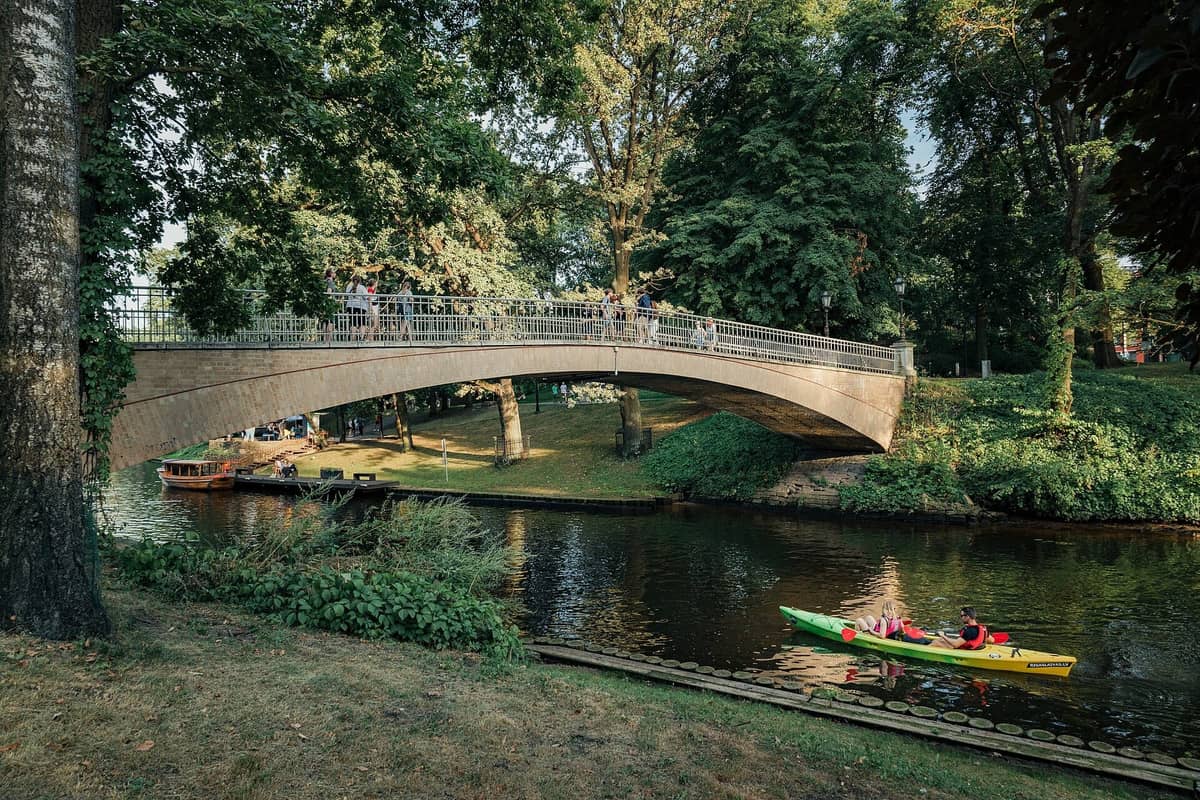 Eine Steinbrücke überquert den Stadtkanal im Bastejkalns-Park, während Menschen darüber gehen und ein Kajak auf dem ruhigen Wasser darunter paddelt.