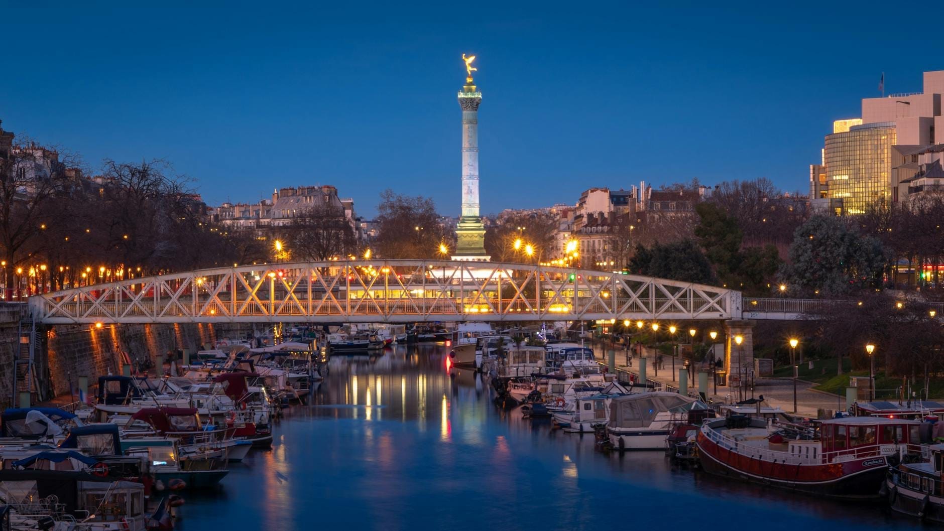 Vista serale della colonna della Bastiglia e del porto turistico con barche ormeggiate lungo l'acqua, un ponte pedonale in primo piano e i riflessi delle luci della città sul canale.