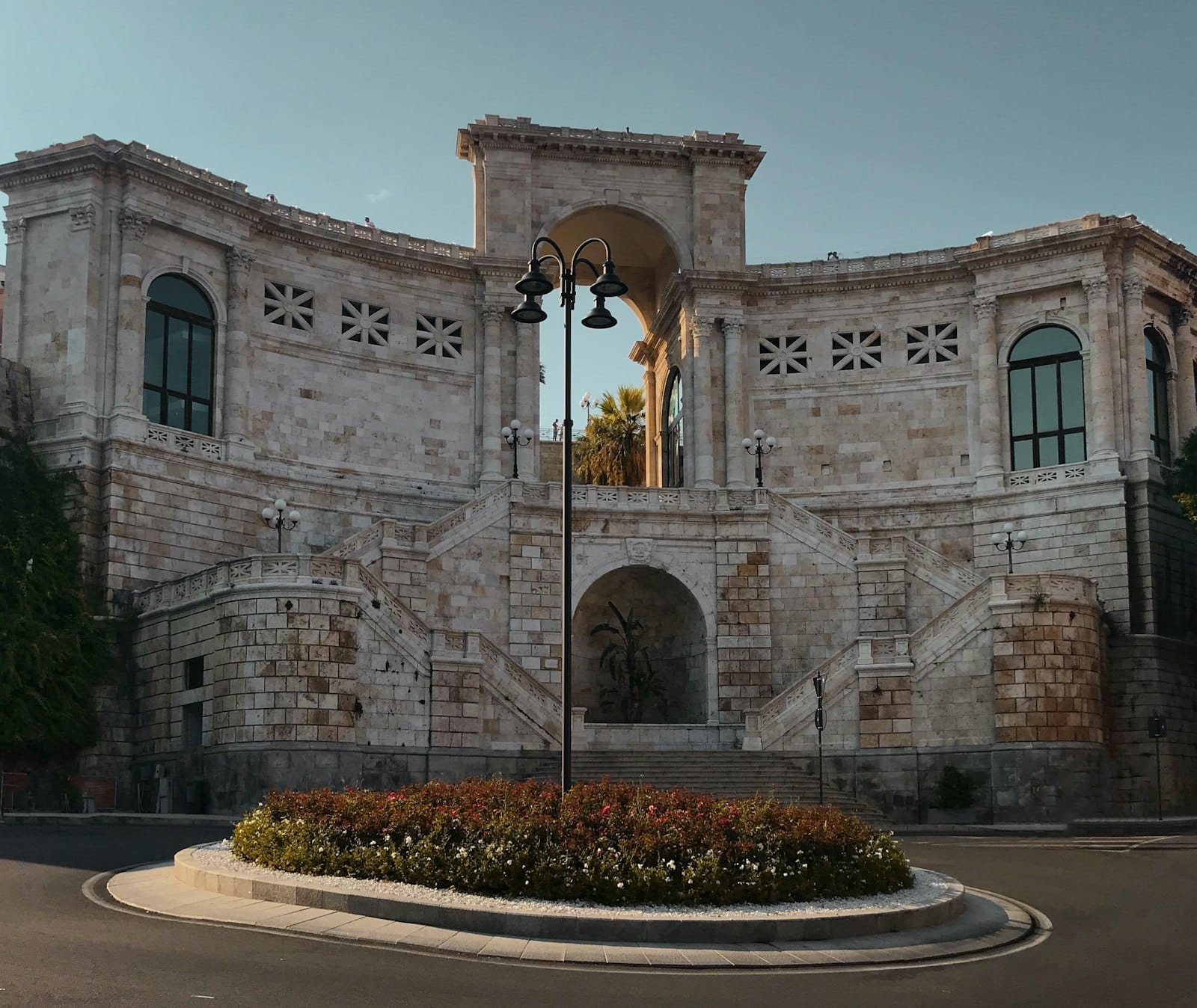 Der Bastione di Saint Remy in Cagliari zeigt beeindruckende weiße Steinbögen, eine doppelte Treppenanlage und einen Kreisverkehr mit Blumen unter einem klaren Himmel.