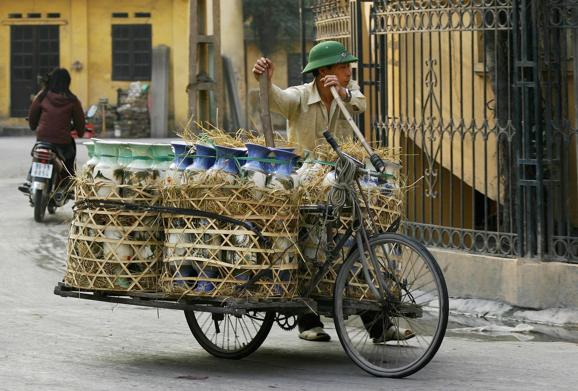 A man transports large painted pottery vases by bicycle cart through the streets of Bát Tràng Ceramic Village, with yellow buildings in the background.