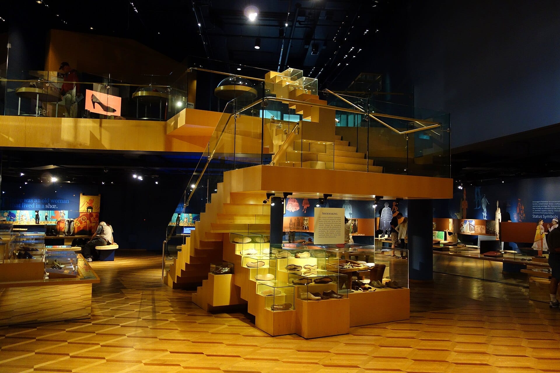 มุมมองภายใน Bata Shoe Museum ที่แสดงให้เห็นบันไดไม้ ตู้แสดงรองเท้าแบบกระจก และผู้เยี่ยมชมที่กำลังสำรวจพื้นที่จัดแสดงที่มีแสงสว่างเพียงพอ