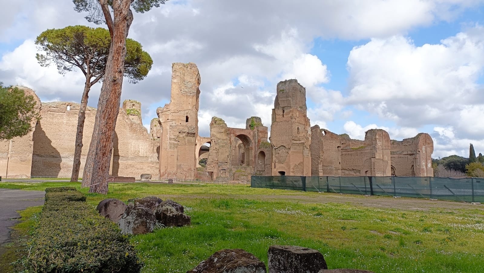 The ruins of the Baths of Caracalla in Rome, with towering ancient brick walls, scattered stones, trees, and a dramatic sky overhead.