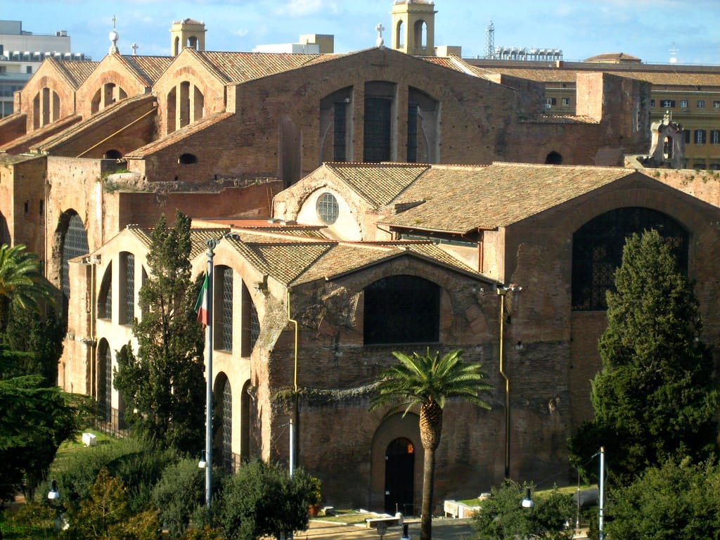The Baths of Diocletian in Rome, featuring massive brick arches, tall windows, and lush greenery under bright daylight.