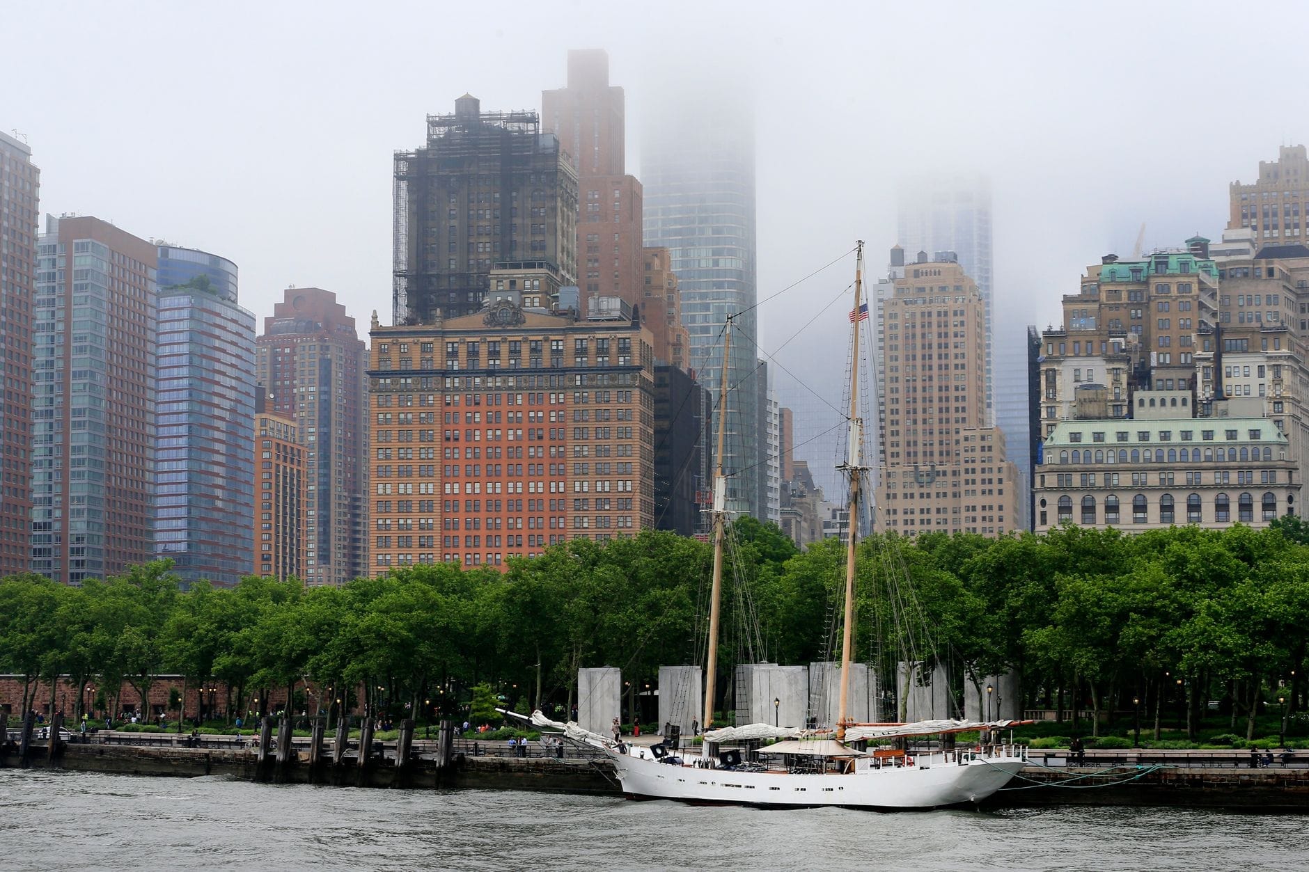 Una barca a vela bianca ormeggiata vicino al lungomare di Battery Park, con alberi rigogliosi e i grattacieli storici di Manhattan che si stagliano tra la foschia sullo sfondo.
