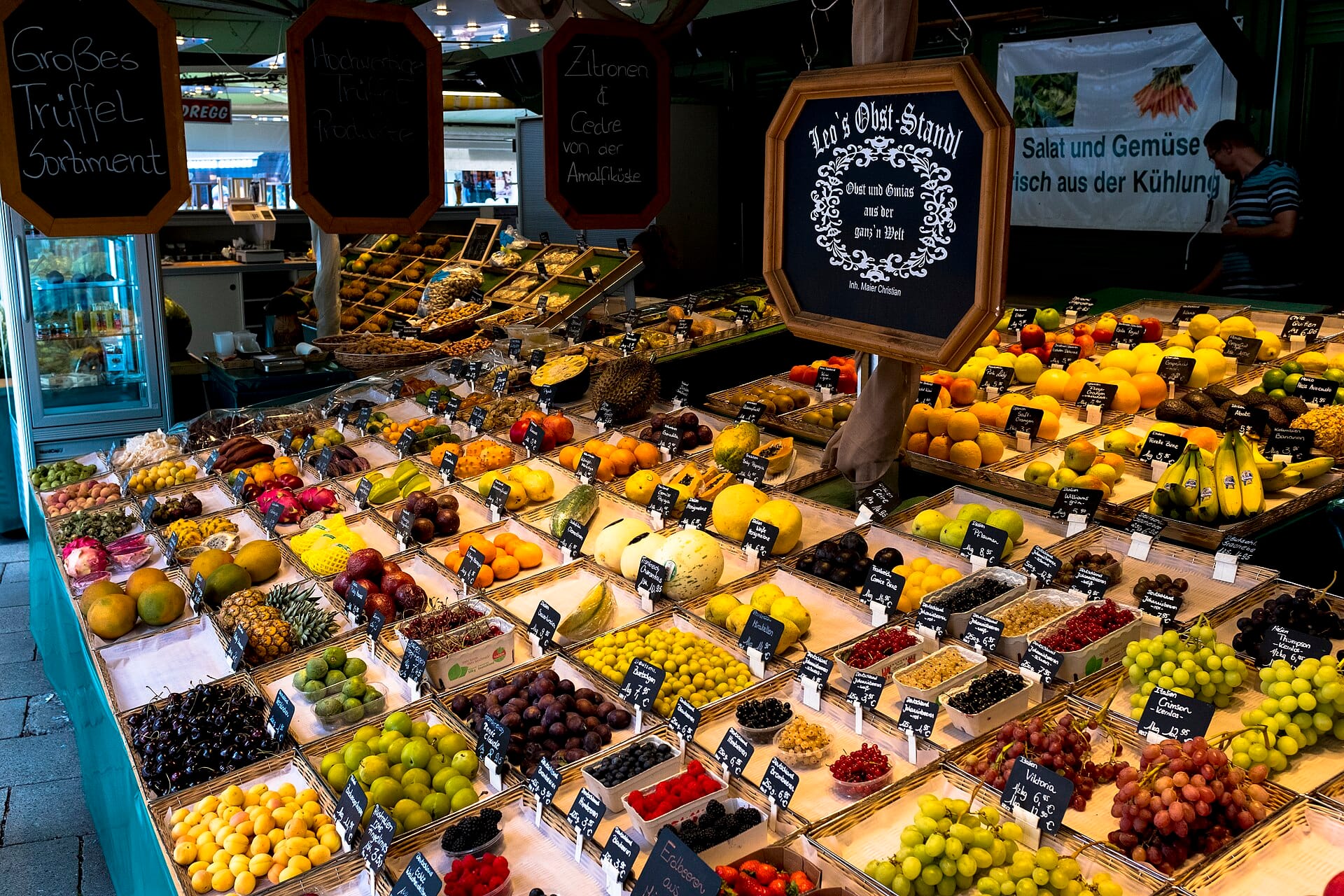แผงผลไม้หลากสีสันพร้อมป้ายภาษาเยอรมันในตลาดในร่ม Viktualienmarkt เมืองมิวนิก นำเสนอผลไม้สดและผลผลิตทางการเกษตรให้เลือกมากมาย