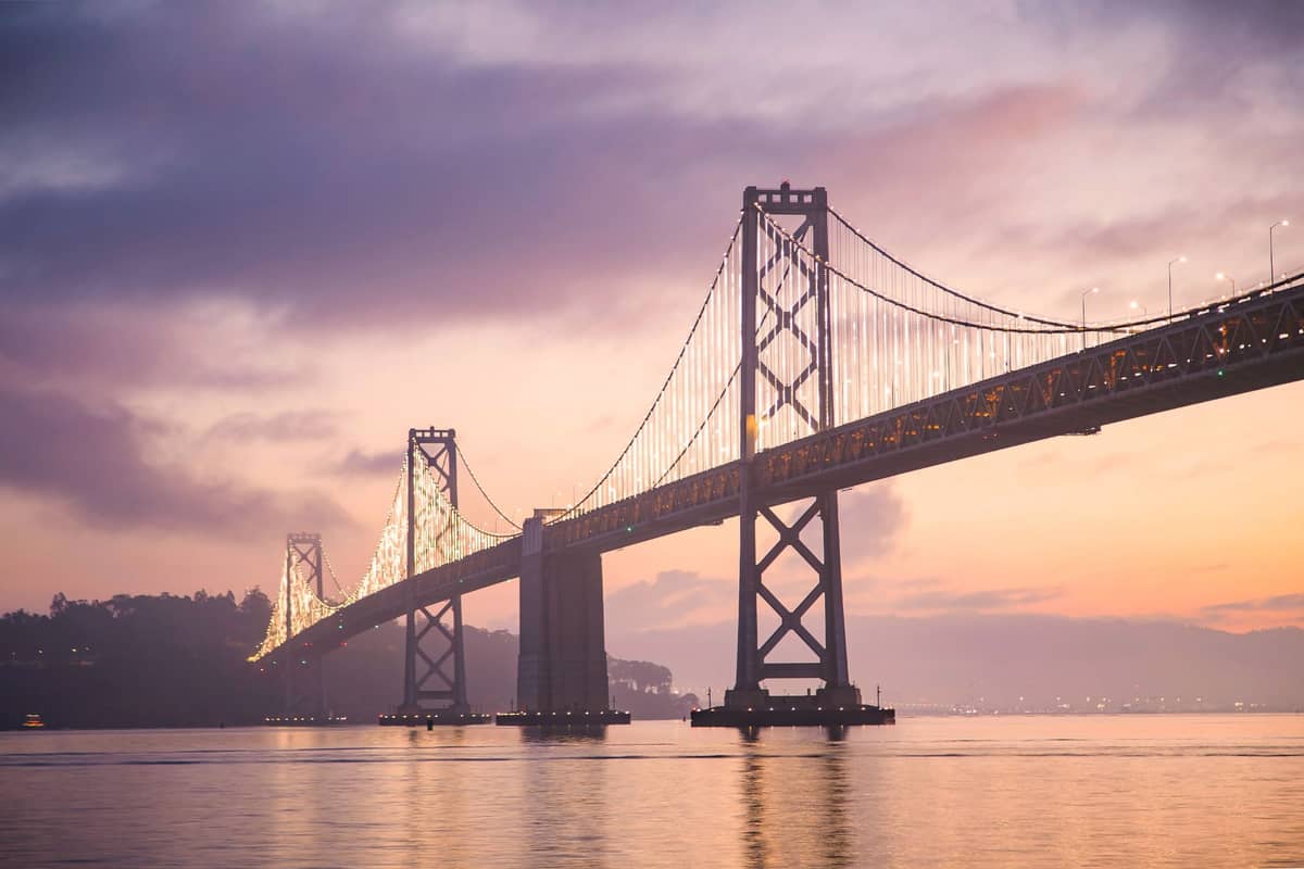 Vista del Bay Bridge San Francisco–Oakland al tramonto, con le luci che si riflettono sull'acqua e un cielo nuvoloso dai colori vividi sullo sfondo.