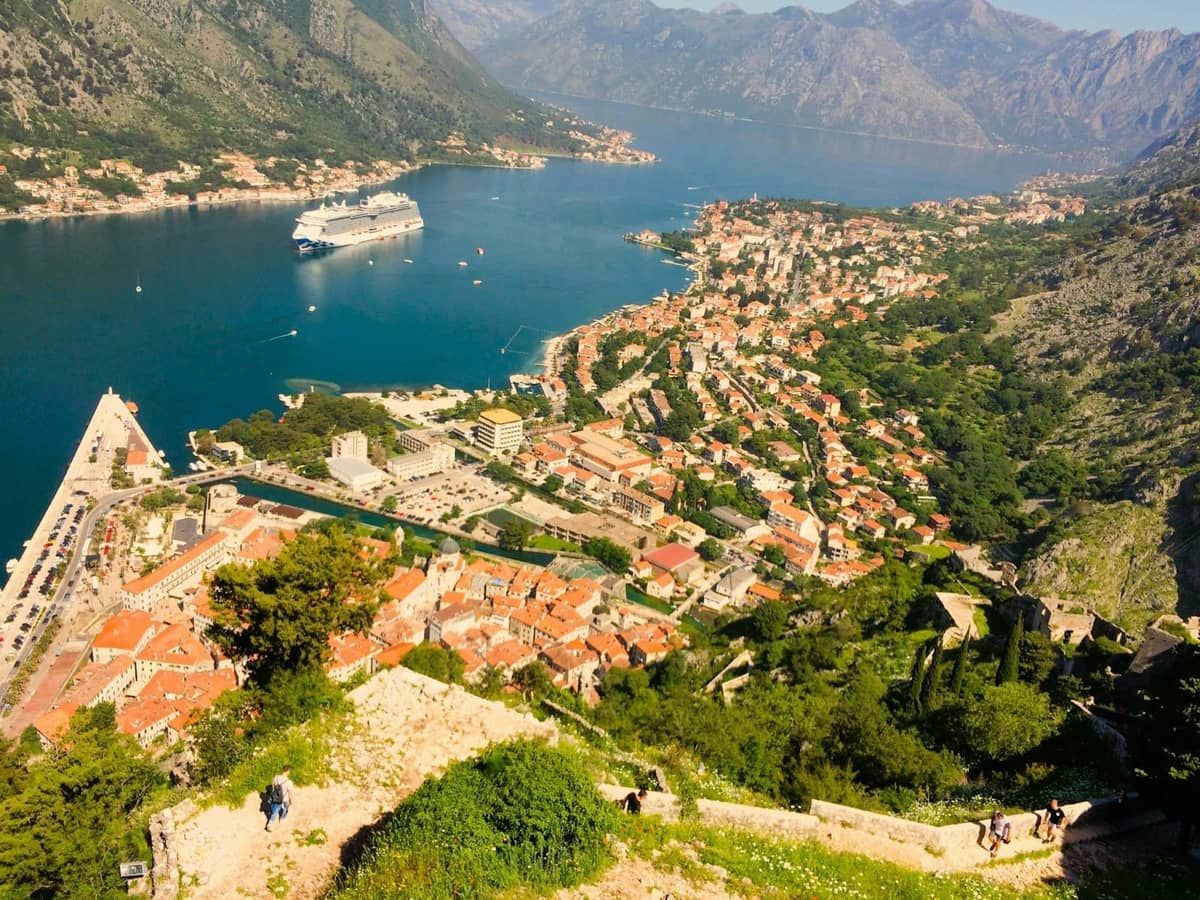 Aerial view of Kotor bay with cruise ship, red-roofed old town, and surrounding green mountains on a sunny day.
