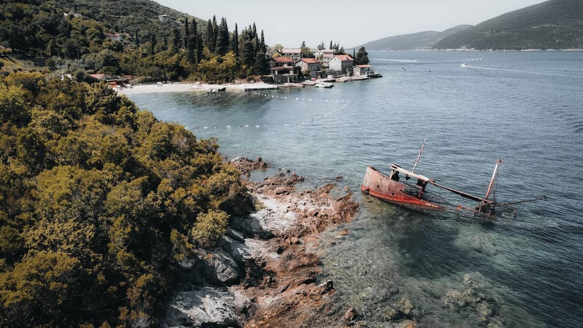 Aerial view of a small bay with a shipwrecked boat near rocky shore and a village in the background along Kotor Bay's coastline.