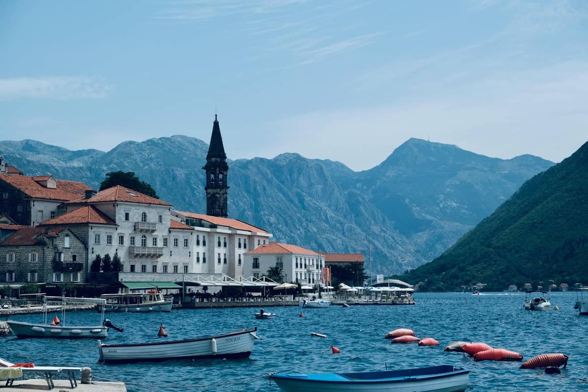 Picturesque waterfront view of Perast in Bay of Kotor, with boats, elegant historic buildings, church tower, and surrounding mountains under a blue sky.