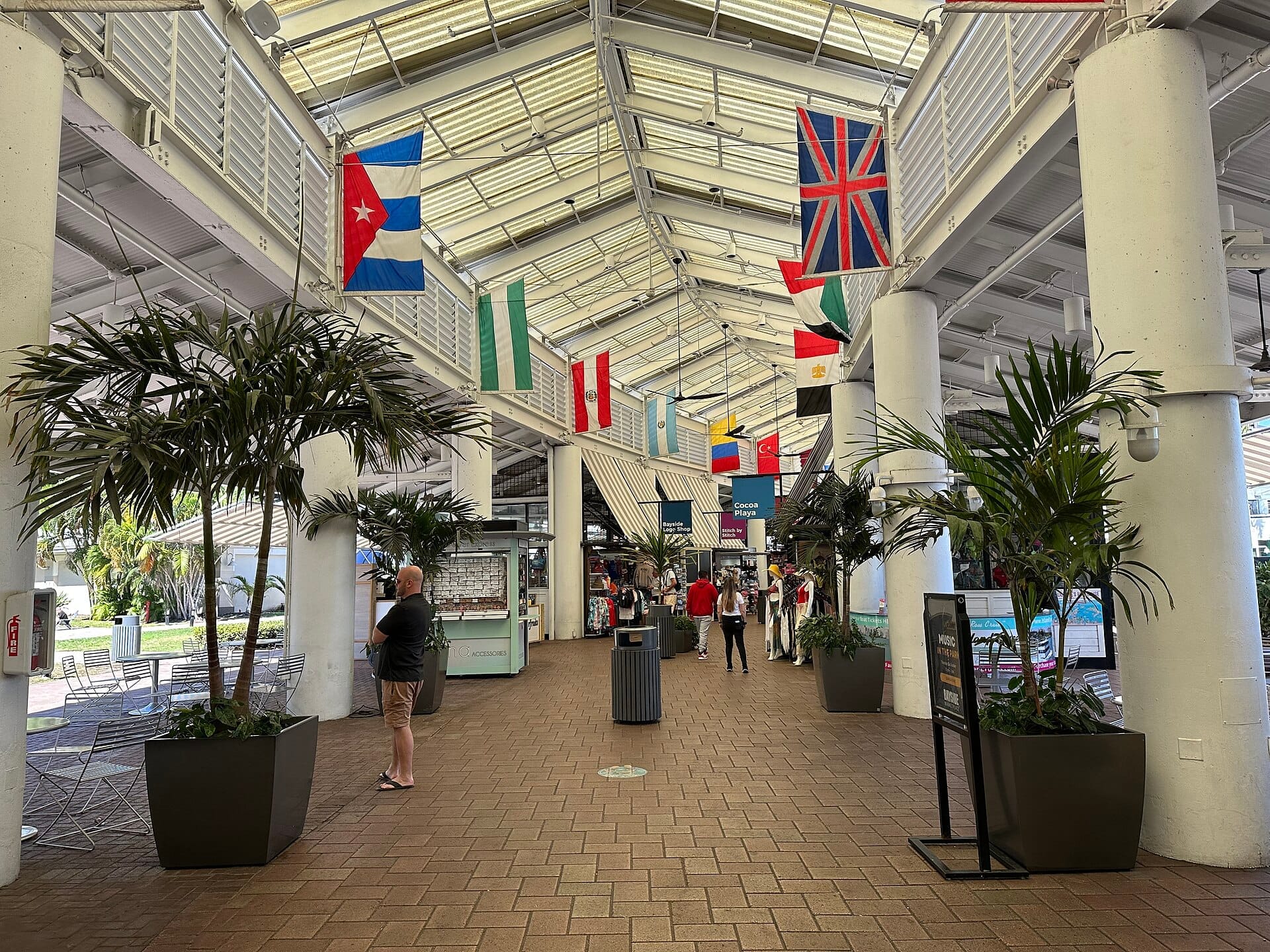 Corridoio interno del Bayside Marketplace con bandiere internazionali, piante tropicali e diverse persone che fanno shopping sotto un luminoso tetto di vetro illuminato dal sole.
