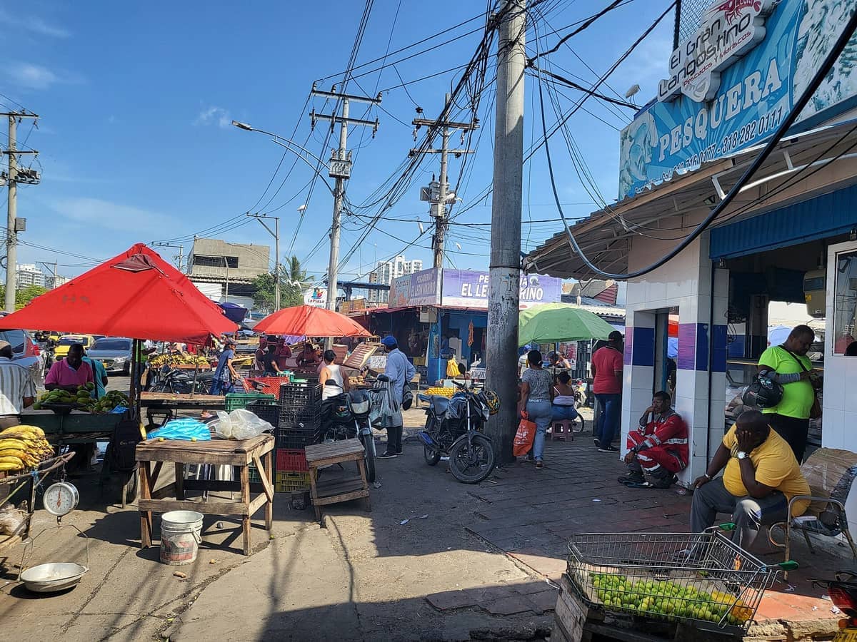 Vendors and shoppers gather under colorful umbrellas at Mercado de Bazurto in Cartagena, surrounded by fresh produce stalls and street activity.