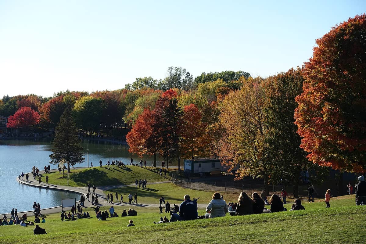 Menschen entspannen und spazieren um den Beaver Lake im Parc du Mont-Royal in Montréal, umgeben von bunten Herbstbäumen und ruhigem Wasser an einem sonnigen Tag.