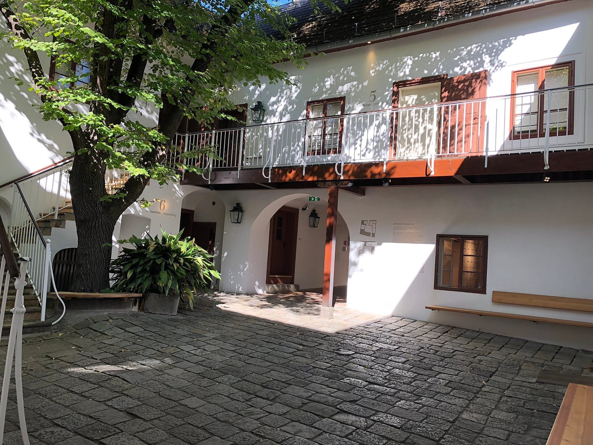 Innenhof des Beethoven Museum Heiligenstadt mit Kopfsteinpflaster, weißen Wänden, Holzbänken und belaubtem Baum im Tageslicht.