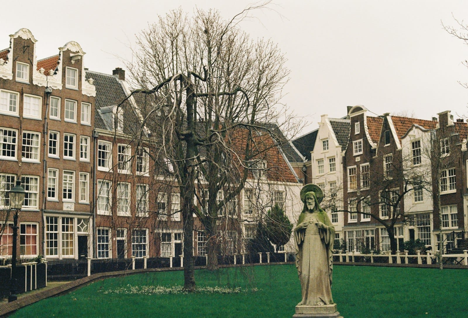 Hilera de casas históricas holandesas y una estatua en el jardín central del Begijnhof Amsterdam, con árboles sin hojas bajo un cielo nublado.