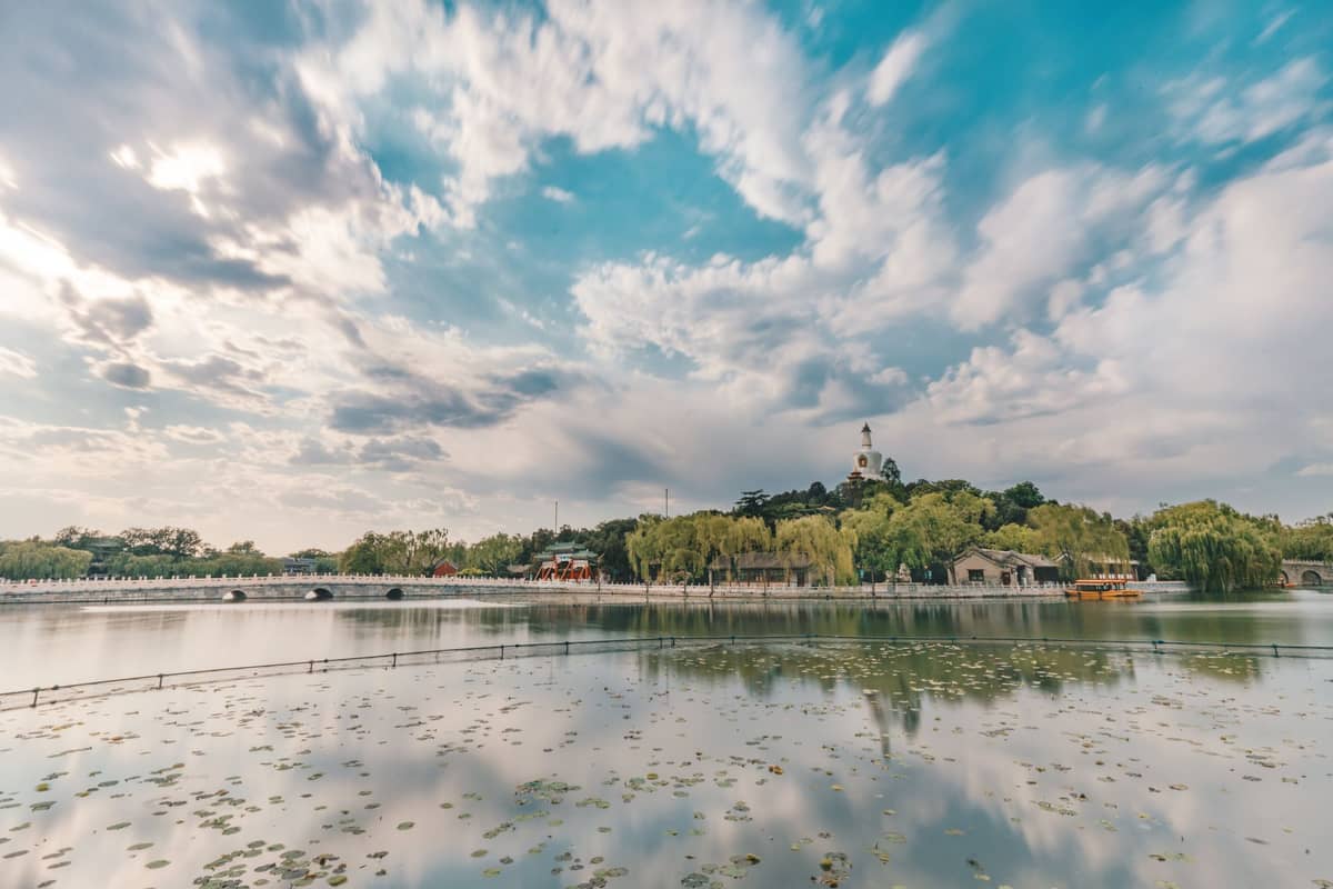 Weitwinkelpanorama des Beihai-Parks mit dem zentralen See, üppigem Grün und der markanten Weißen Dagoba auf dem Hügel unter einem dramatischen Himmel.