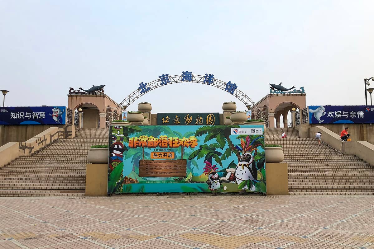 Eingang des Pekinger Aquariums mit seinem markanten Torbogen, Meerestierstatuen und bunten Werbebannern an der Haupttreppe.