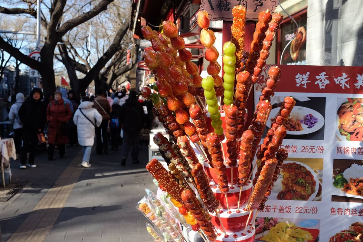 Brochettes d'aubépine enrobées de sucre aux couleurs vives exposées sur un stand de street food animé avec des personnes marchant en arrière-plan en pleine journée.