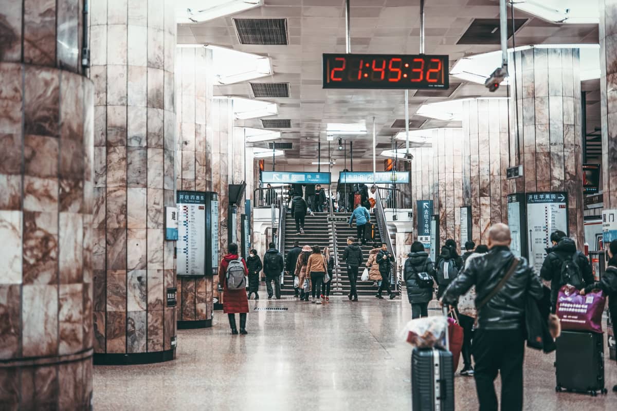 Des voyageurs avec des bagages et des navetteurs circulent dans une station de métro ou de train aux murs en marbre à Beijing, avec une grande horloge numérique en hauteur et des panneaux en mandarin.