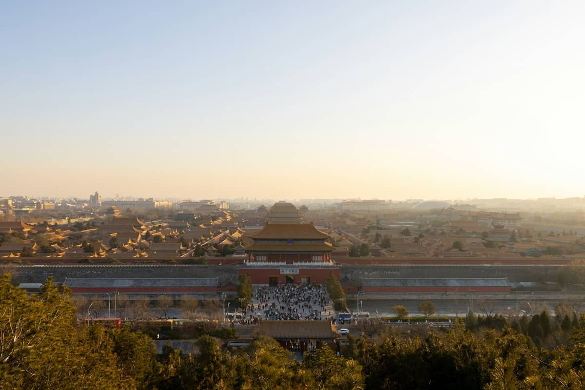 Vue large de la Cité Interdite à Pékin au coucher du soleil avec des personnes rassemblées à son entrée et les toits brillant d'une lumière chaleureuse.