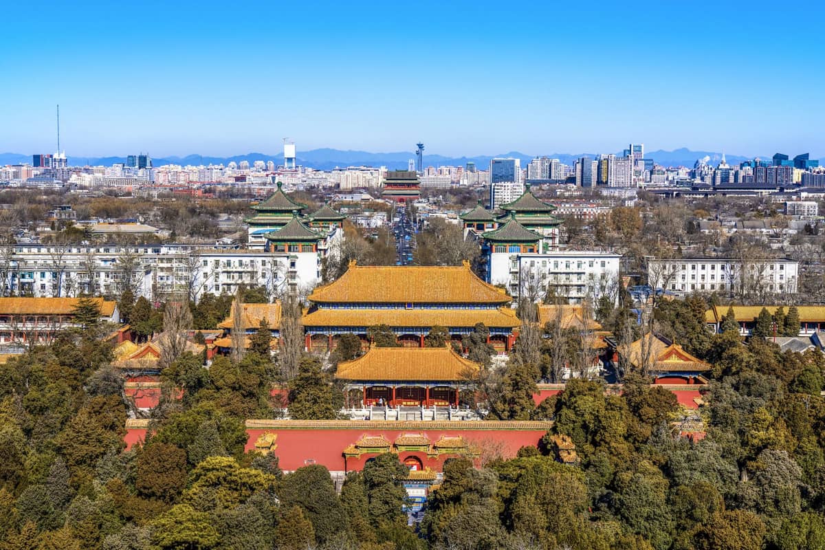 A clear wide view of Beijing’s Forbidden City surrounded by green trees, with the city skyline and mountains visible in the background under a bright blue sky.