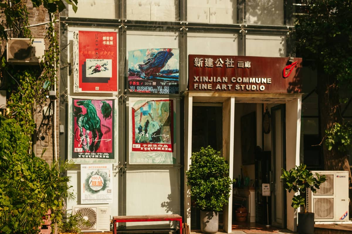 Eingang zu einem zeitgenössischen Kunstatelier in Pekings 798 Art Zone – das Schild zeigt XinJian Commune Fine Art Studio, umgeben von bunten Ausstellungsplakaten und Grünpflanzen.