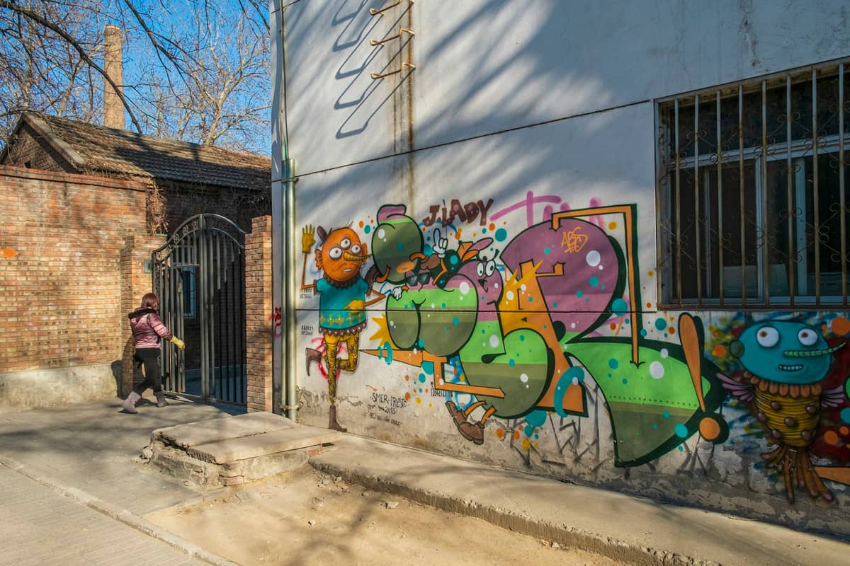 Eine Straße im Kunstbezirk 798 in Peking mit bunten Graffiti an einer Wand und einer gehenden Person, Bäume werfen Herbstschatten.