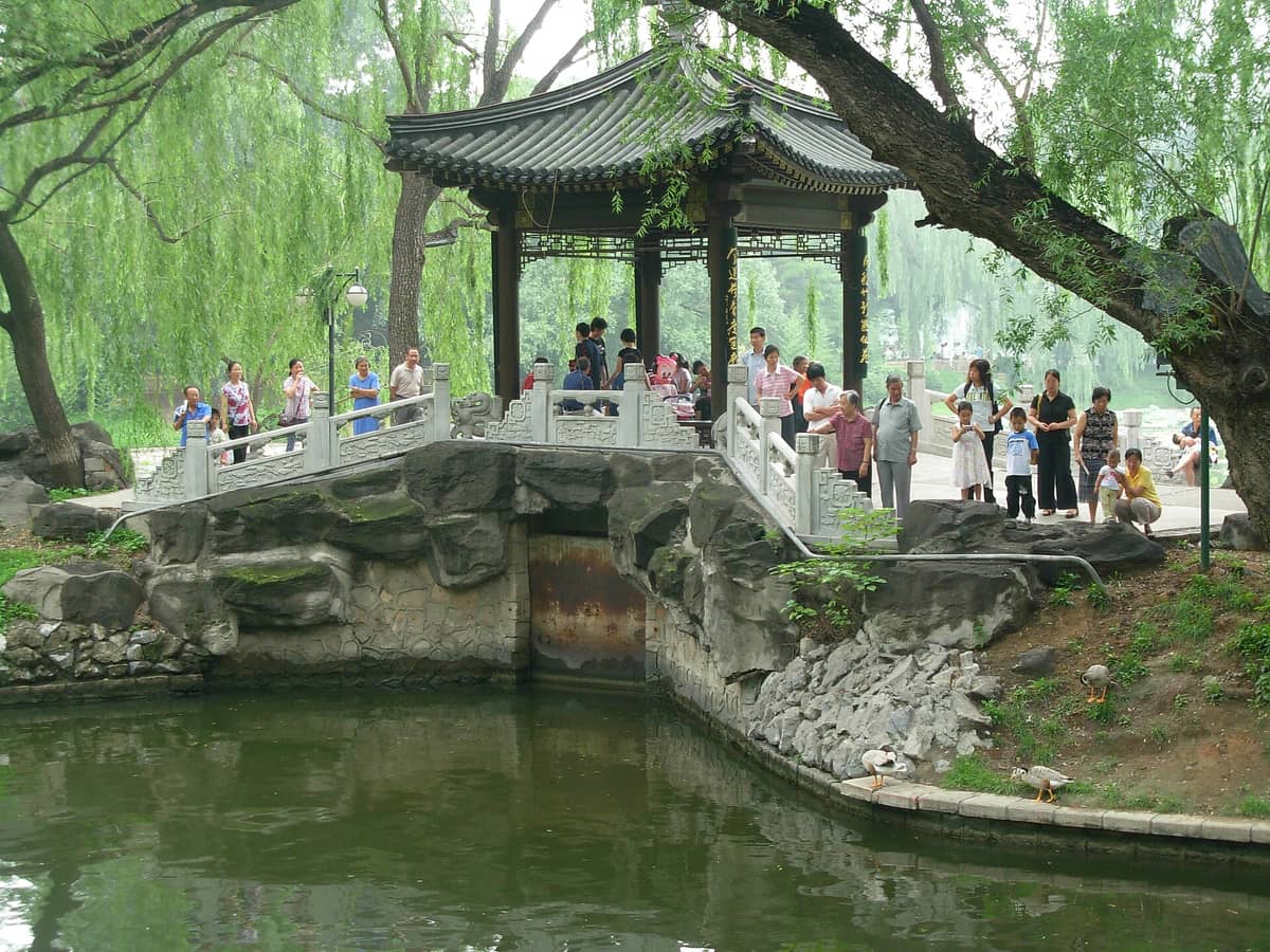 Menschen versammeln sich in einem üppigen traditionellen chinesischen Pavillon an einem von Weiden umgebenen Teich in einem Park in Beijing.