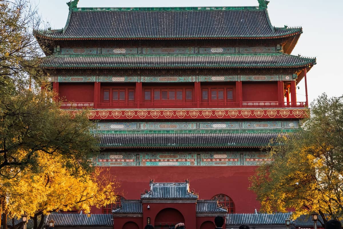 Der Beijing Drum Tower, ein großes rotes historisches Gebäude mit grünem Ziegeldach, Bäume im Vordergrund, an einem klaren Tag.
