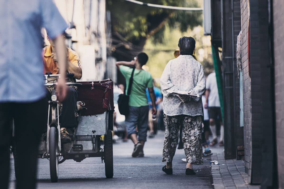Belebte Hutong-Szene in Beijing mit Fußgängern und einem Rickshaw-Pedicab, das tagsüber durch eine schmale Gasse fährt.