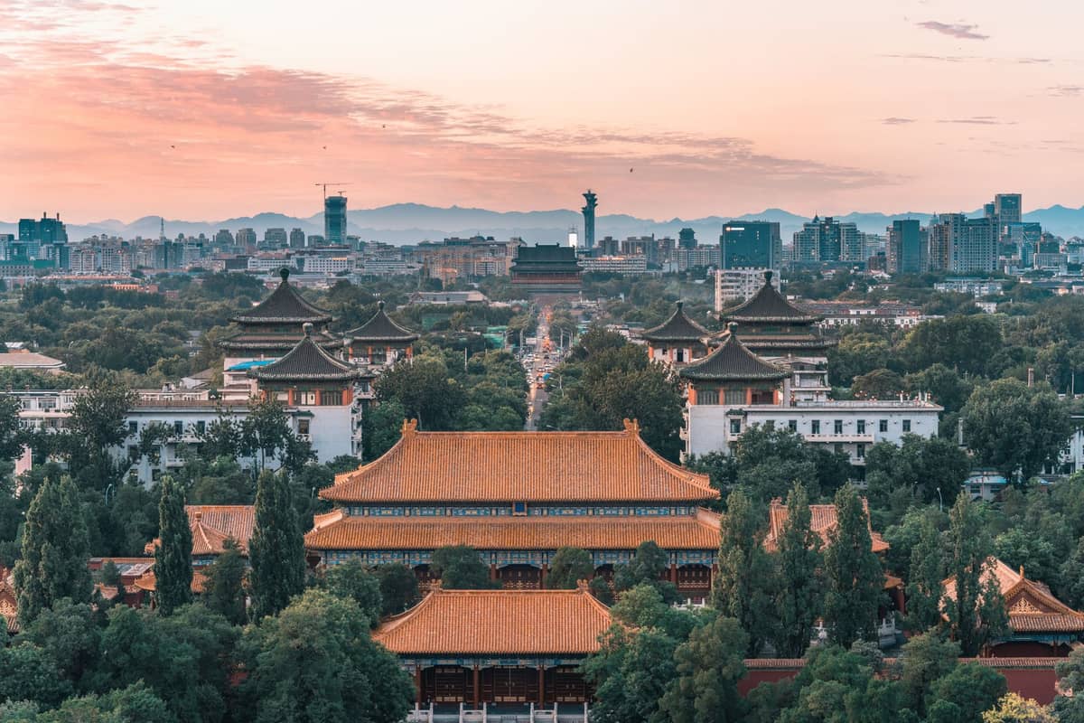 Vue panoramique du centre de Pékin montrant des toits traditionnels de l'ère impériale, des avenues bordées d'arbres et des immeubles lointains avec des montagnes à l'horizon.