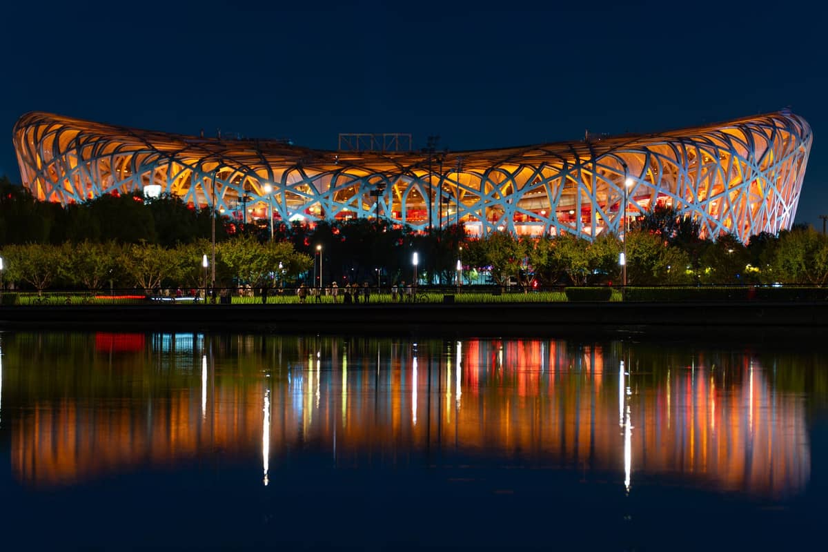 Nachtansicht des Nationalen Olympiastadions Peking (Vogelnest), hell erleuchtet und im ruhigen Wasser davor gespiegelt.