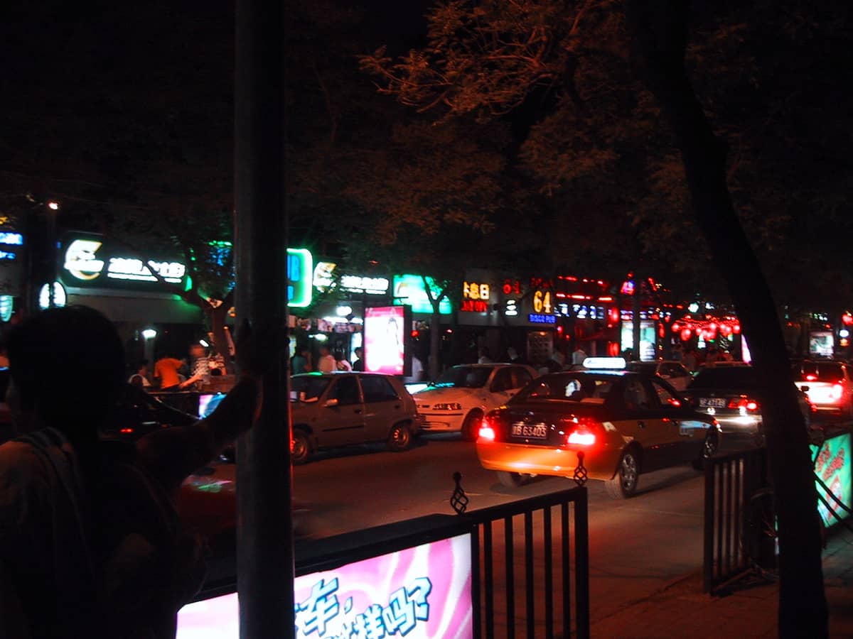 Nächtliche Straßenszene in Beijing mit Bars, Neonlichtern und Taxis, die vor belebten Nachtclubs auf Fahrgäste warten.