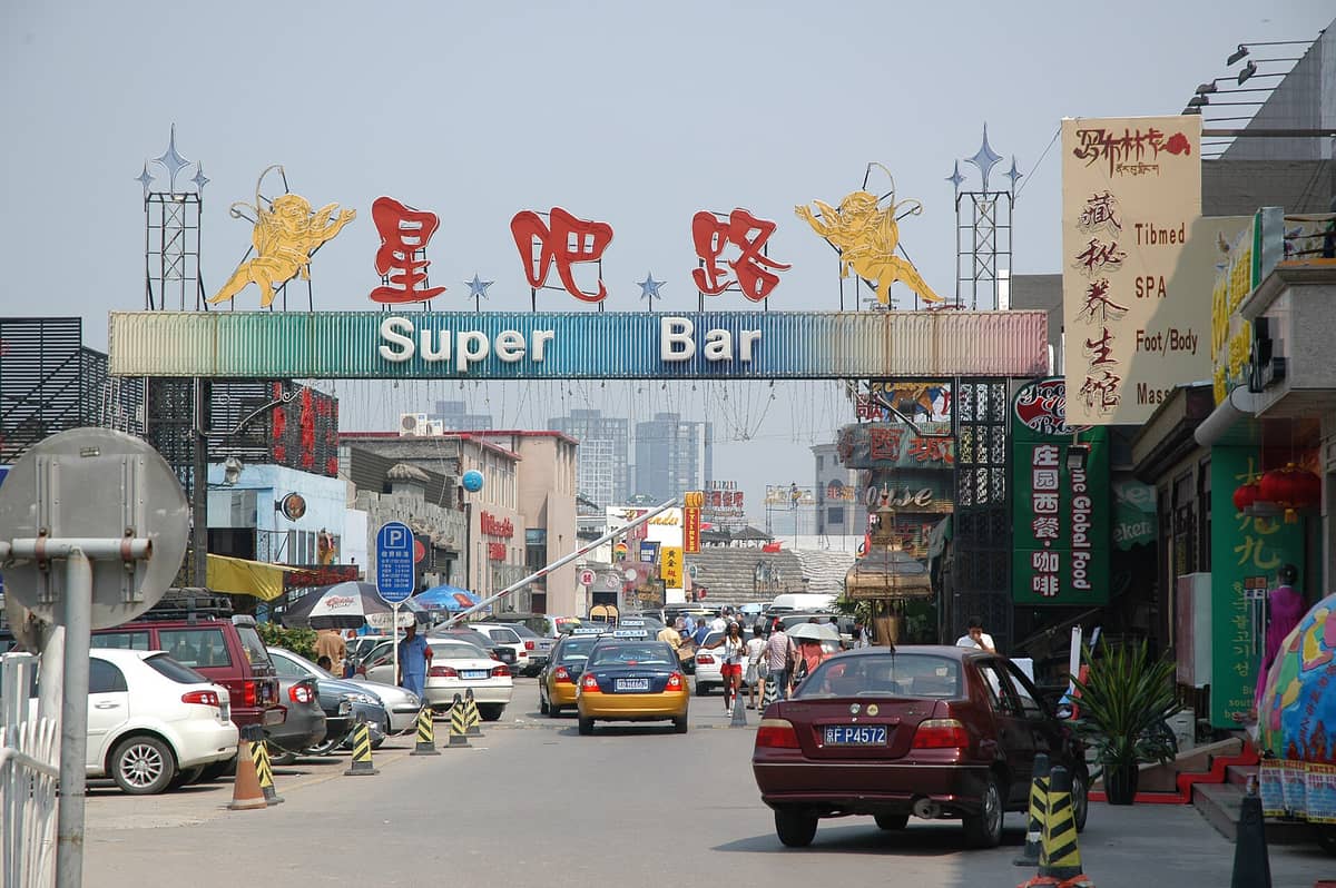 Straßenszene am Eingang der berühmten Sanlitun Bar Street in Beijing mit dem Schild 'Super Bar', tagsüber belebt von Autos und Menschen.