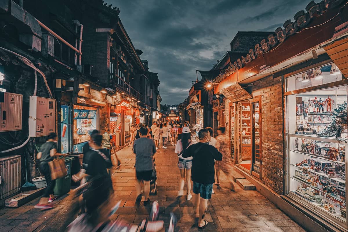 Belebte Fußgängerzone in Beijing in der Abenddämmerung, gesäumt von traditionellen Geschäften und leuchtenden Lichtern, mit Menschen, die spazieren und einkaufen.