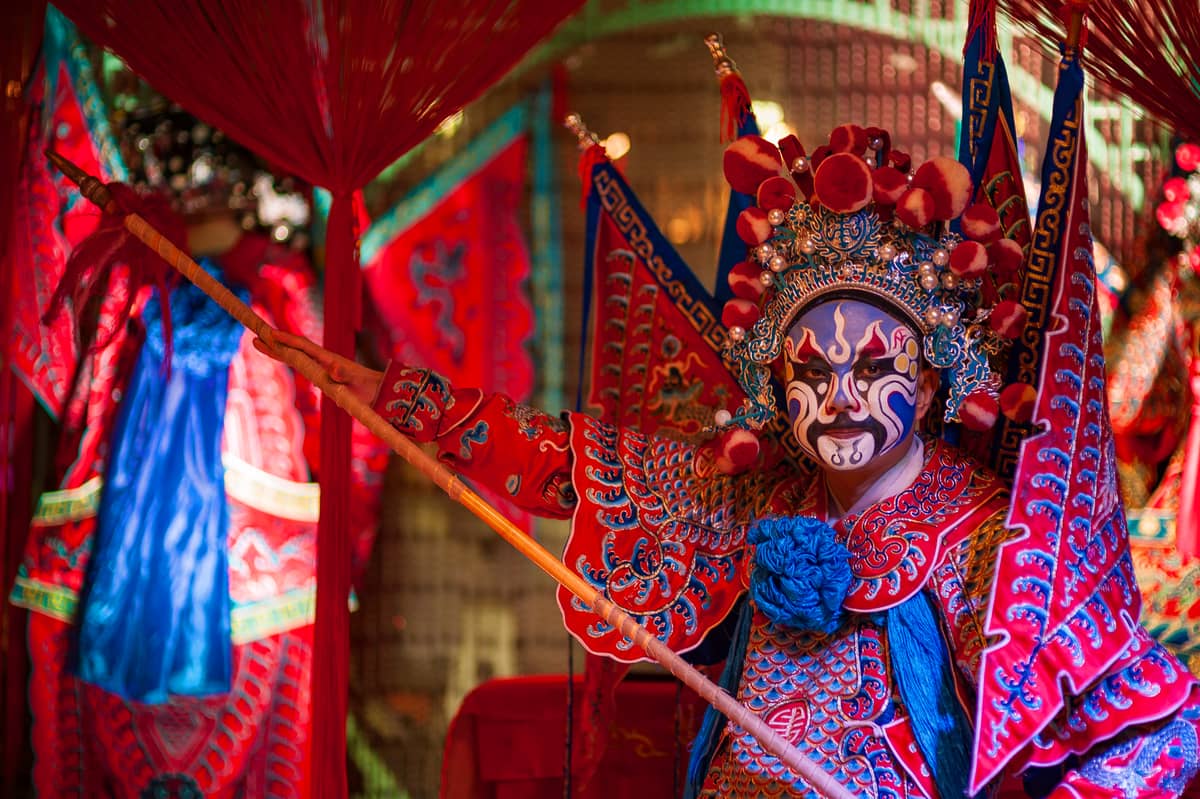 Peking-Oper-Darsteller in aufwendigem roten Kostüm und traditioneller Gesichtsbemalung, einen Stab haltend, umgeben von farbenfrohen Bühnenrequisiten und Kostümen.