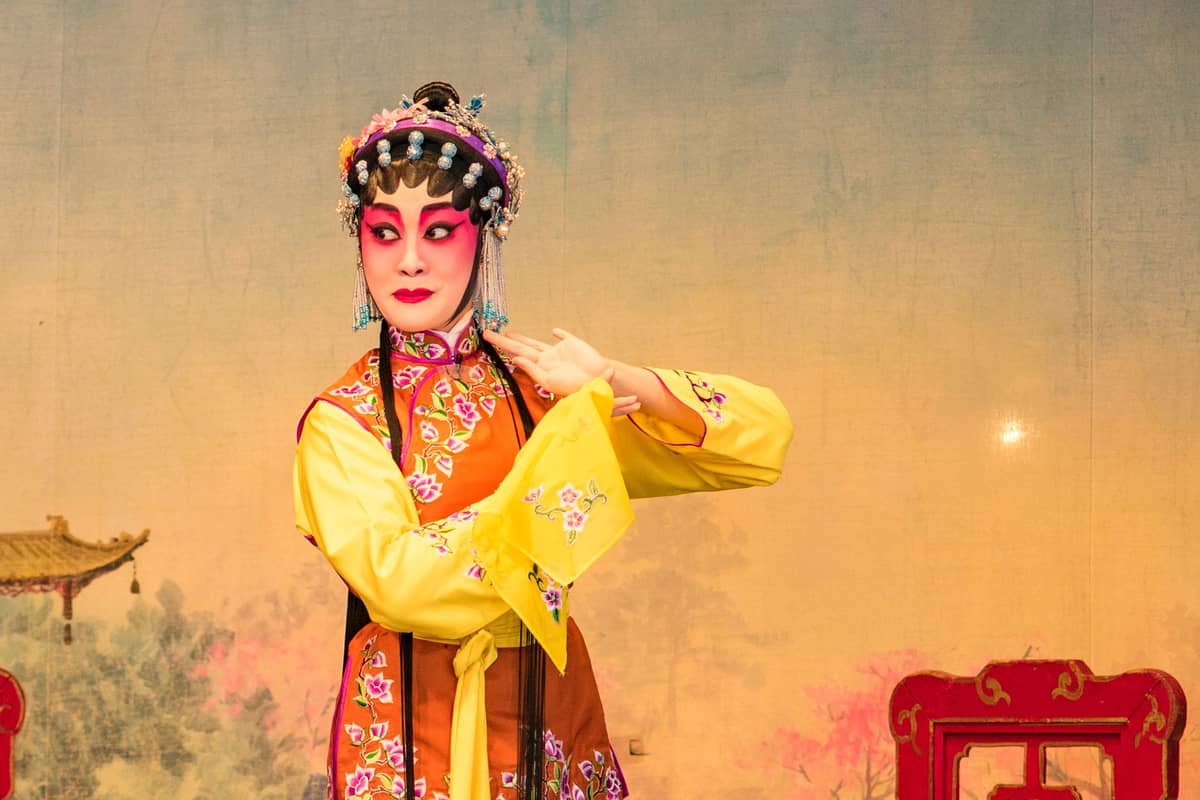 Eine Darstellerin der Pekingoper in traditionellem Kostüm und Bühnenmaske, die in einer dramatischen Pose auf der Bühne steht – vor einem Hintergrund, der an ein klassisches chinesisches Gemälde erinnert.