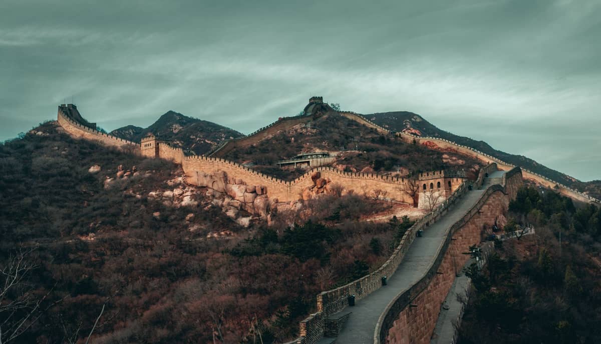 Wide view of the Great Wall of China winding over rugged, mountainous terrain at sunset, with dramatic clouds and warm light covering the stone ramparts.