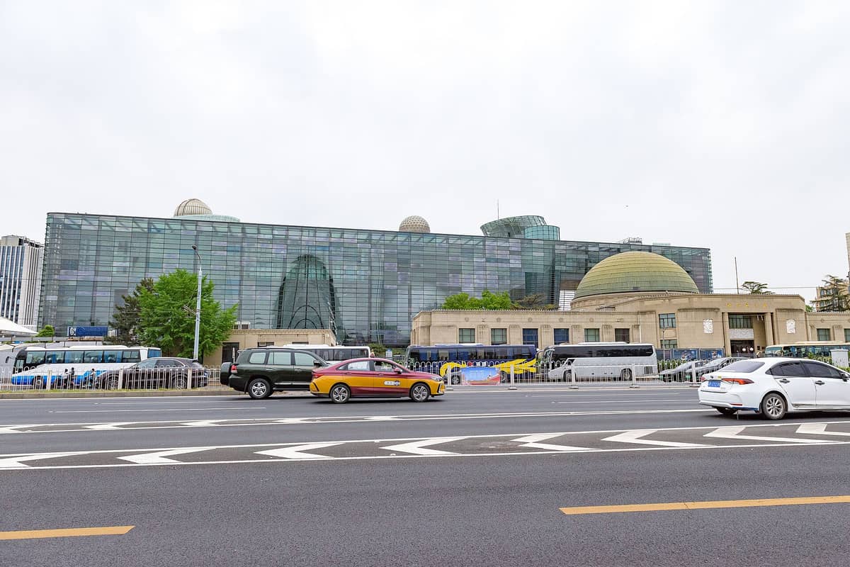Weitwinkelaufnahme der Außenansicht des Pekinger Planetariums mit dem originalen Kuppelbau und dem modernen Glasanbau sowie Autos und Bussen auf der Straße davor.
