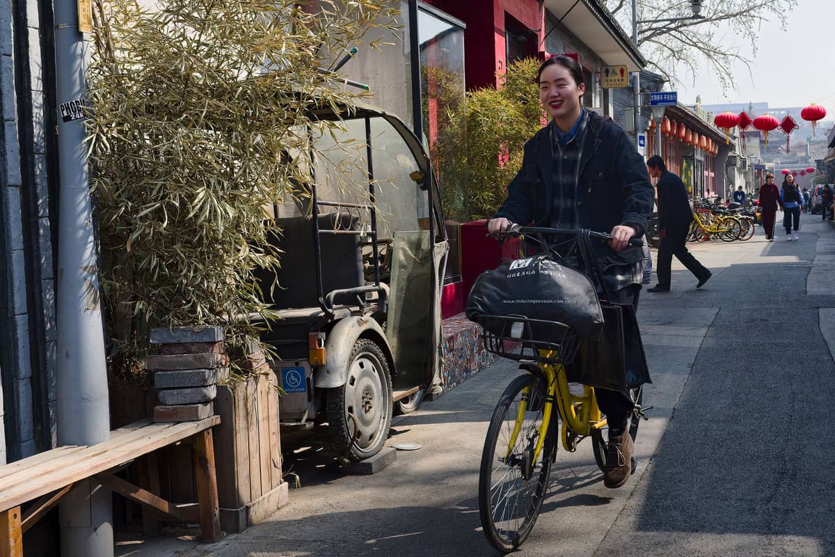 Person auf einem Fahrrad in einer traditionellen Pekinger Hutong-Gasse, gesäumt von Läden, Laternen und charakteristischen grauen Ziegelmauern der Innenhofhäuser.