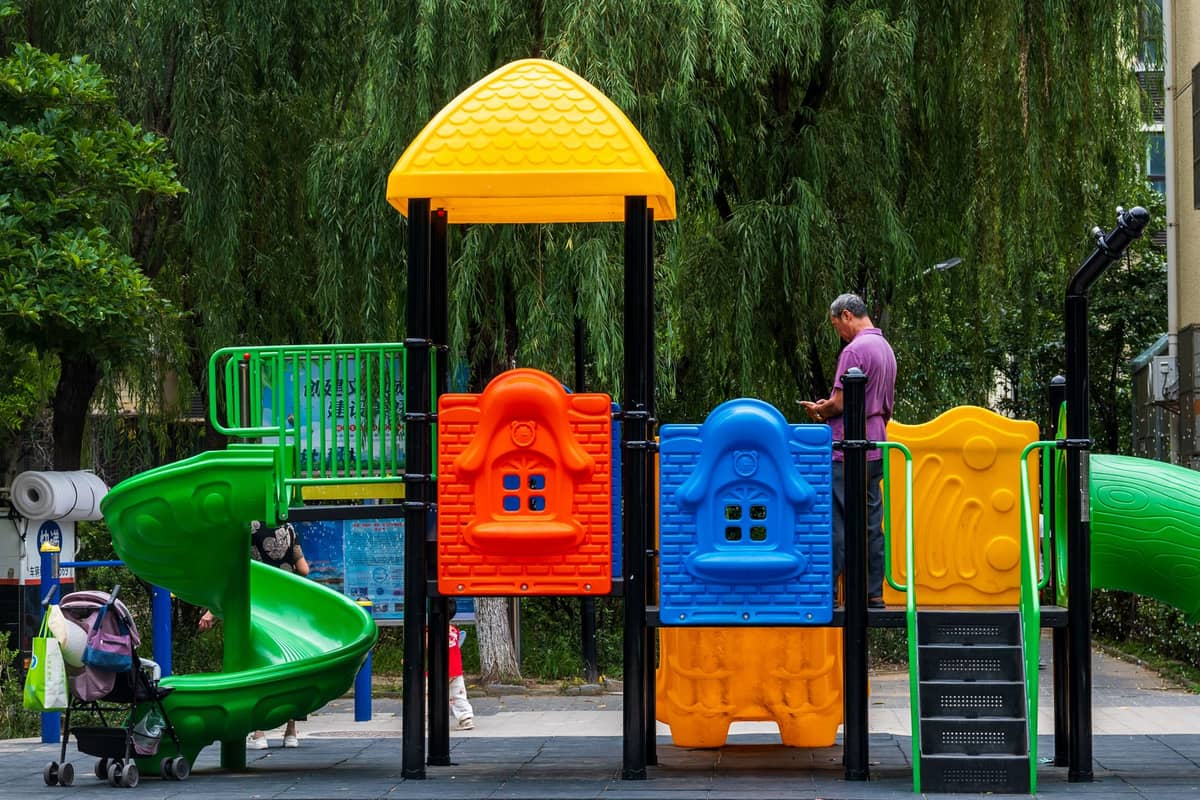 Colorful children's playground with slides in front of trees and urban buildings; stroller and adult visible nearby on a sunny day.