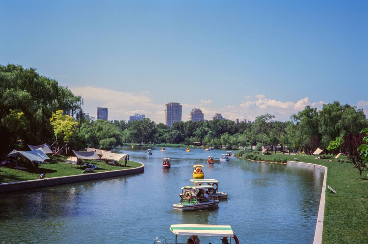 Familien und Kinder auf bunten Tretbooten auf einem See in einem großen Stadtpark in Peking, mit Bäumen und Stadtgebäuden im Hintergrund.