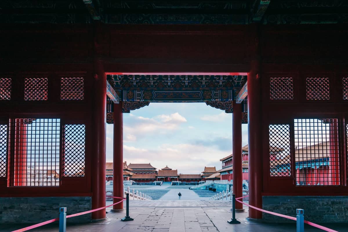 View from inside a red gate looking outward to Forbidden City courtyards, with classical Chinese roofs and a person walking in the distance.