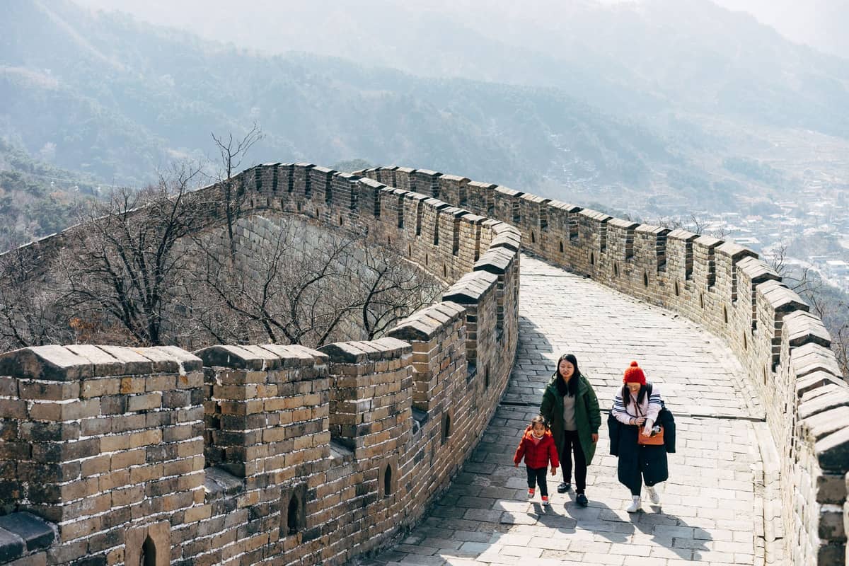 Three people, including two children, walk along the Great Wall of China with scenic mountains in the background.
