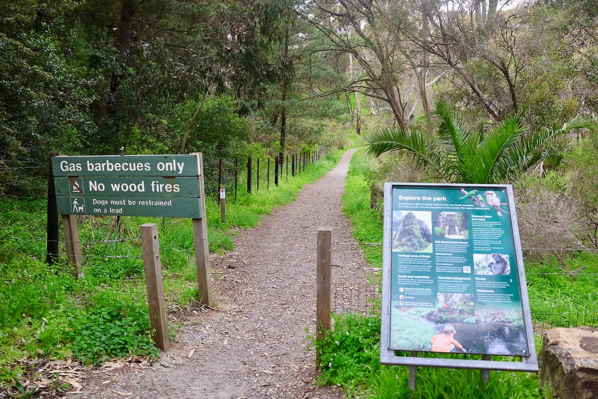 Entrance to a trail at Belair National Park with green bushland, a park rules sign for barbecues and dogs, and an interpretive information board.