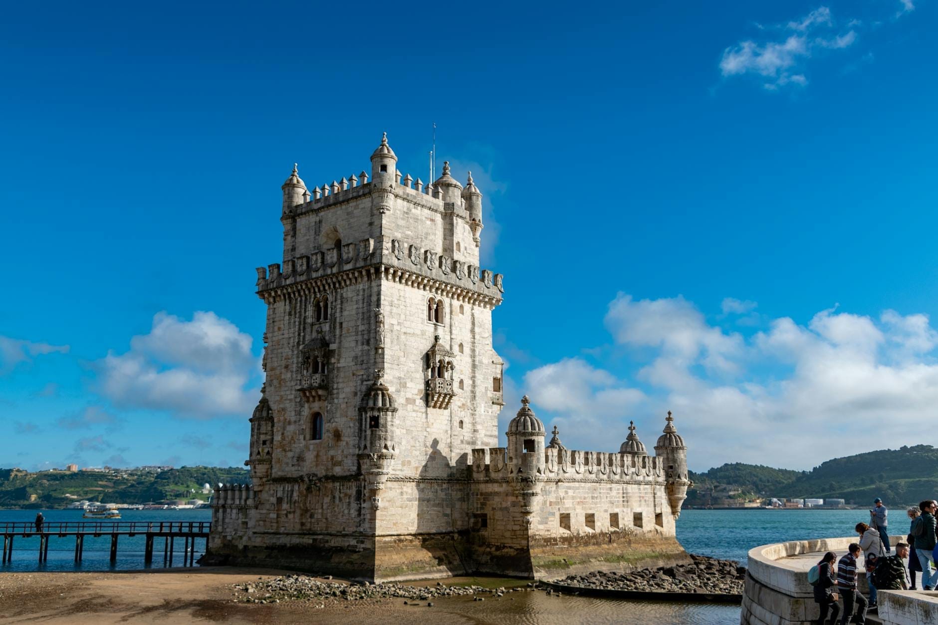 La Torre di Belém si erge accanto al Tago in una giornata di sole, con cielo azzurro e nuvole, visitatori che passeggiano nelle vicinanze e Lisbona sullo sfondo.
