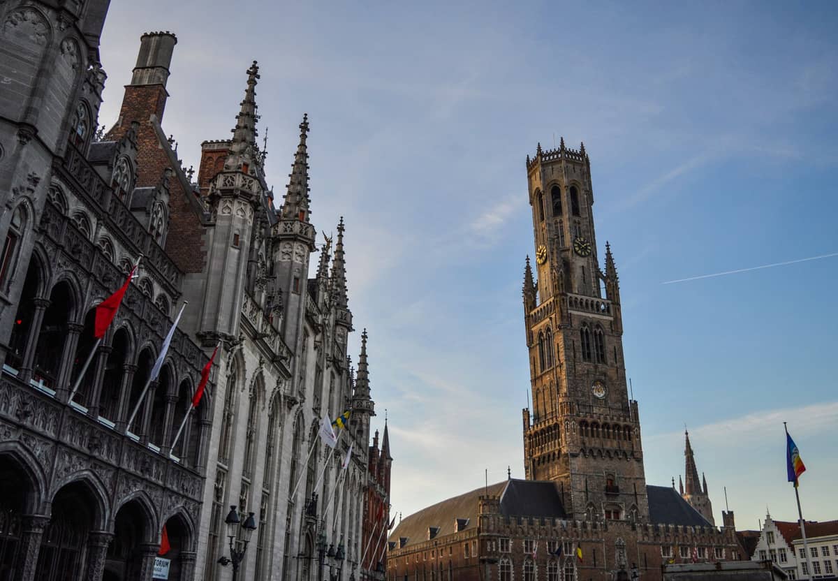 The Belfry of Bruges stands tall over the bustling Markt square, surrounded by historic buildings under a blue sky near sunset.